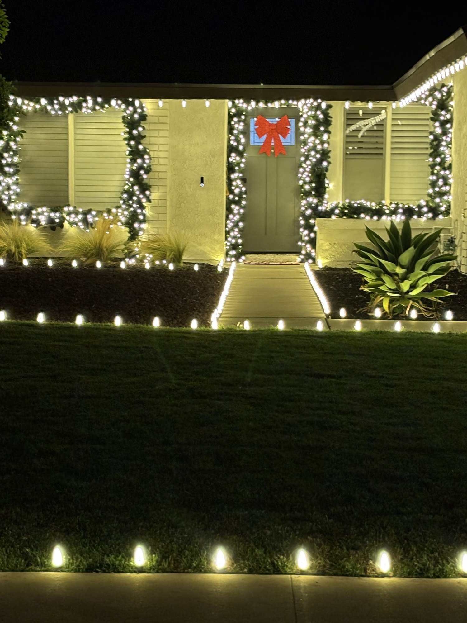 Front door framed in lit garland with a red bow, windows trimmed in lights and a walkway lined with bulbs