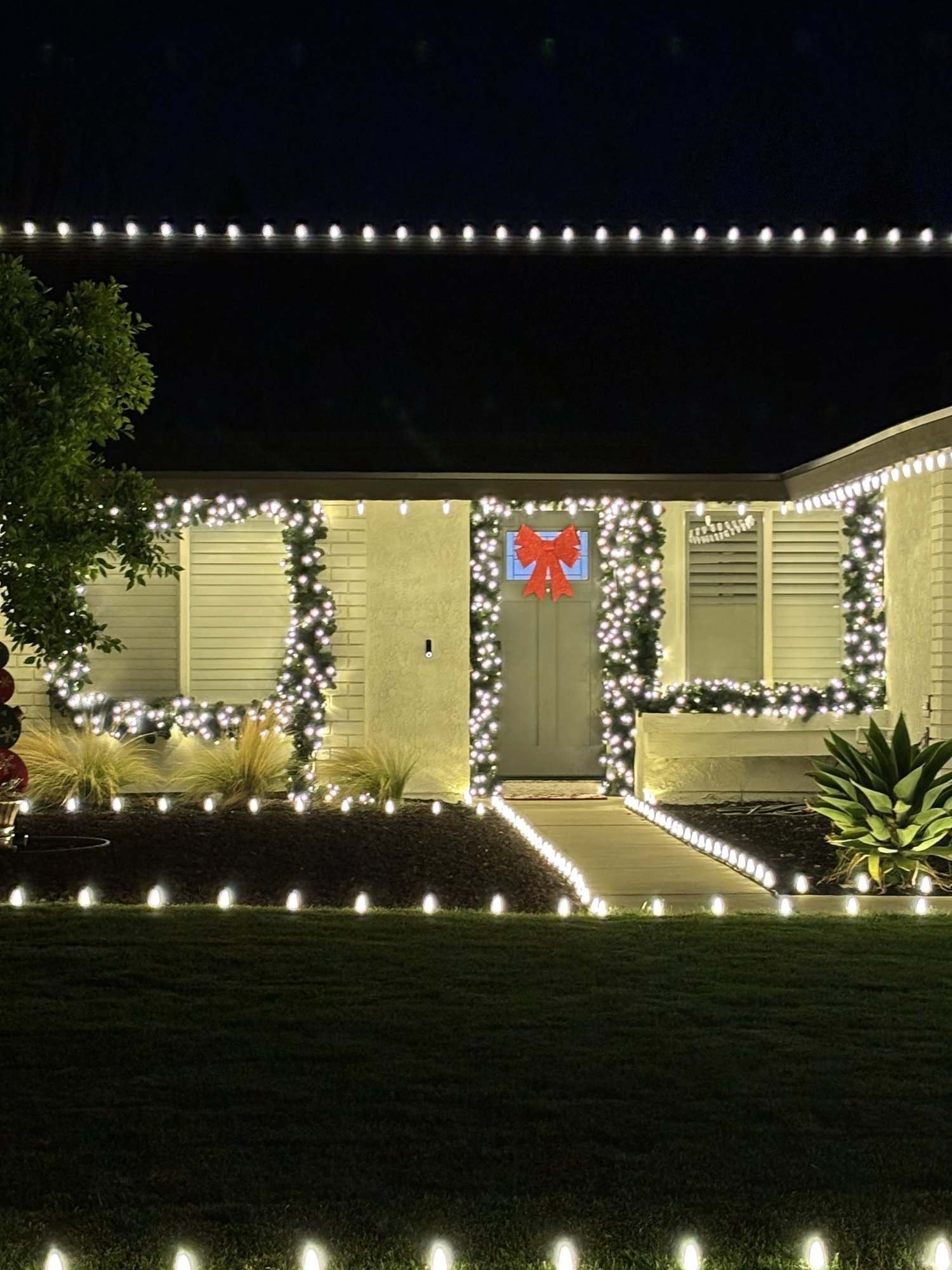 Home with warm white roofline lights, lit garland around the windows and door, a red bow and lighted lawn stakes