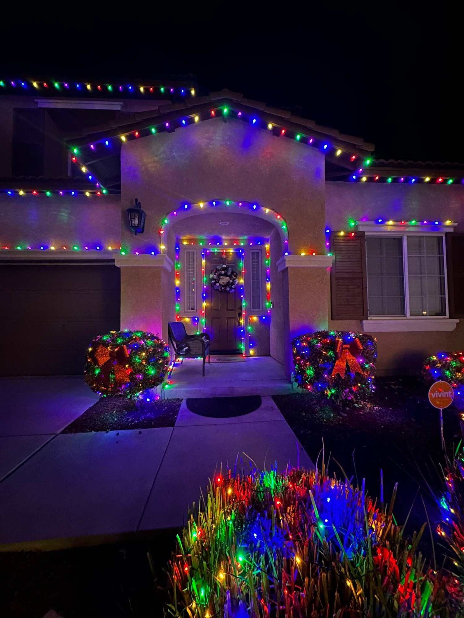 Home with multicolor roofline lights, a lit entry arch and colorful lighted shrubs